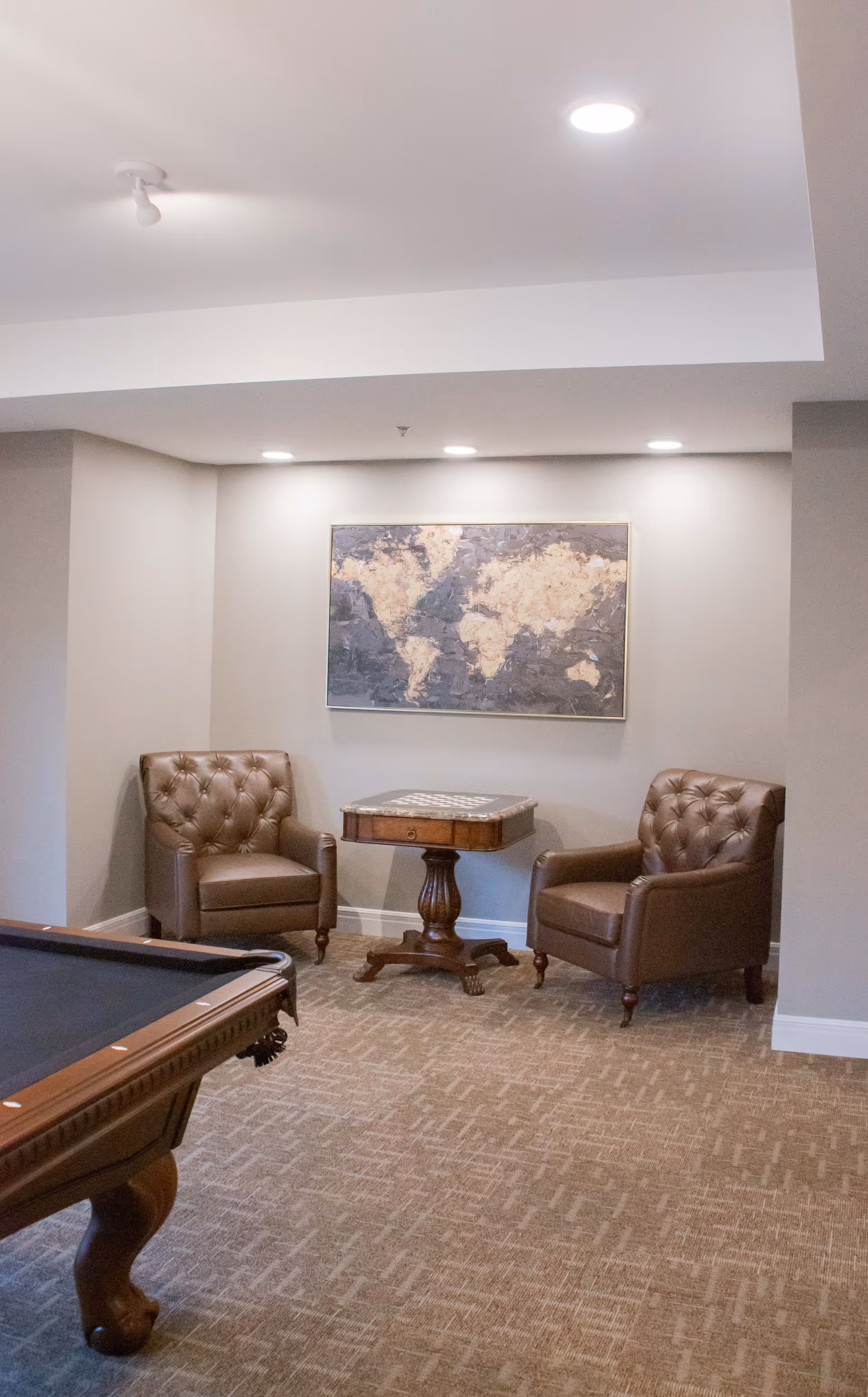 A cozy seating area with two brown leather tufted armchairs and a small wooden table with a chessboard on top, set against a light gray wall with a framed world map artwork. Part of a pool table is visible in the foreground.