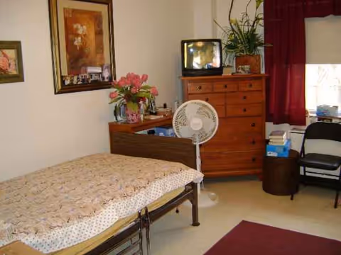 A furnished bedroom with a bed, dresser, fan, TV, plants, and chairs in a senior home.