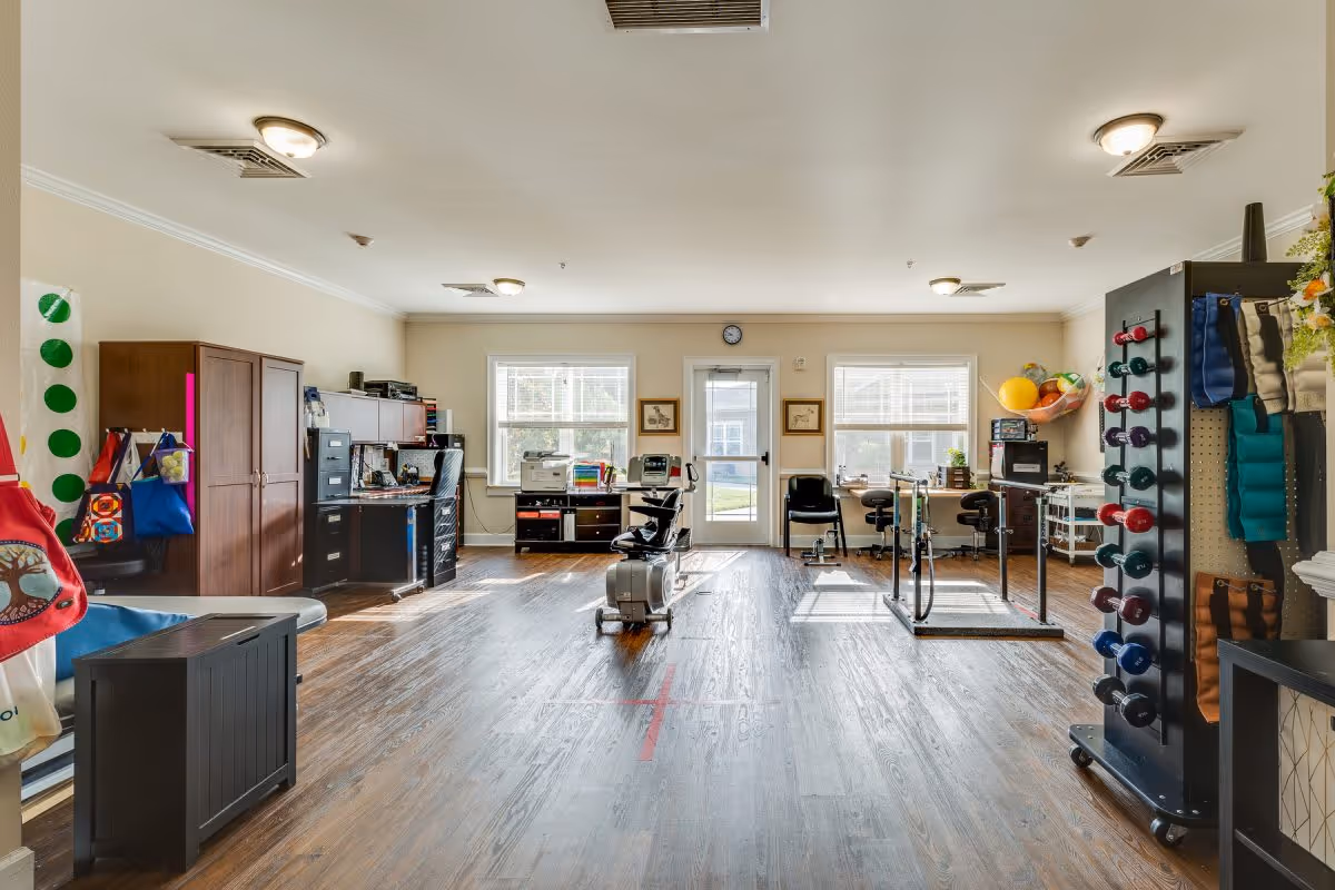 A spacious room with wooden flooring and beige walls, featuring exercise equipment including a stationary bike and parallel bars. There are two windows and a door at the far end letting in natural light. The room also contains office furniture such as desks, chairs, filing cabinets, and shelves with various items. On the right side, there is a rack holding colorful dumbbells and exercise bands. The room appears to be a physical therapy or rehabilitation area within an assisted living facility.