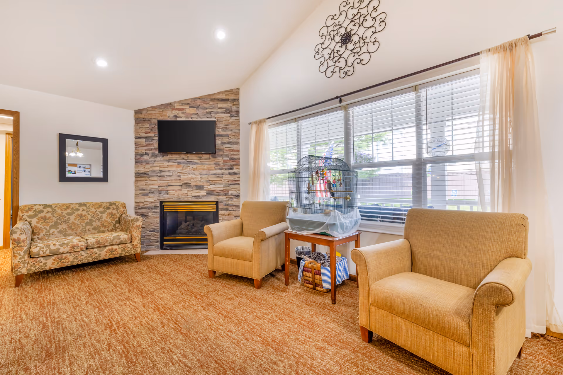 A cozy living room with a floral patterned sofa, two beige armchairs, a stone fireplace with a mounted flat-screen TV above it, and a large window with sheer curtains. A birdcage sits on a small wooden table near the window, and a decorative wall piece hangs above the window.