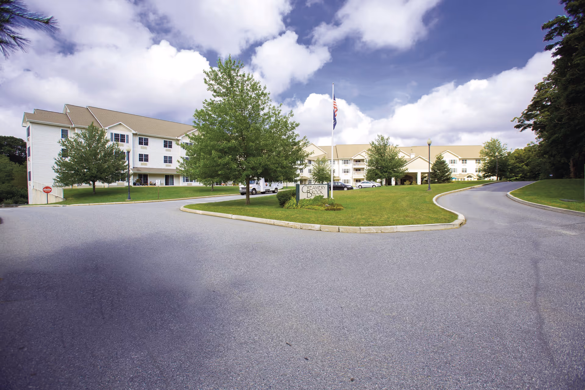 Front exterior of the Creamery Brook Village senior living building with a circular driveway, flagpole, and landscaped lawn.