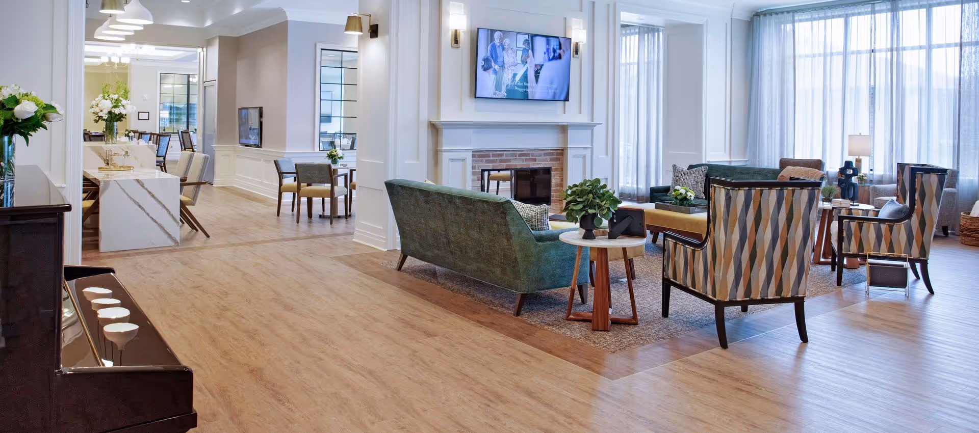 A bright and spacious senior living facility common area with a green sofa, patterned armchairs, a fireplace with a mounted TV above it, and large windows with sheer curtains allowing natural light. There is a piano on the left side and a dining area visible in the background with tables and chairs. The flooring is wood with a carpeted seating area.