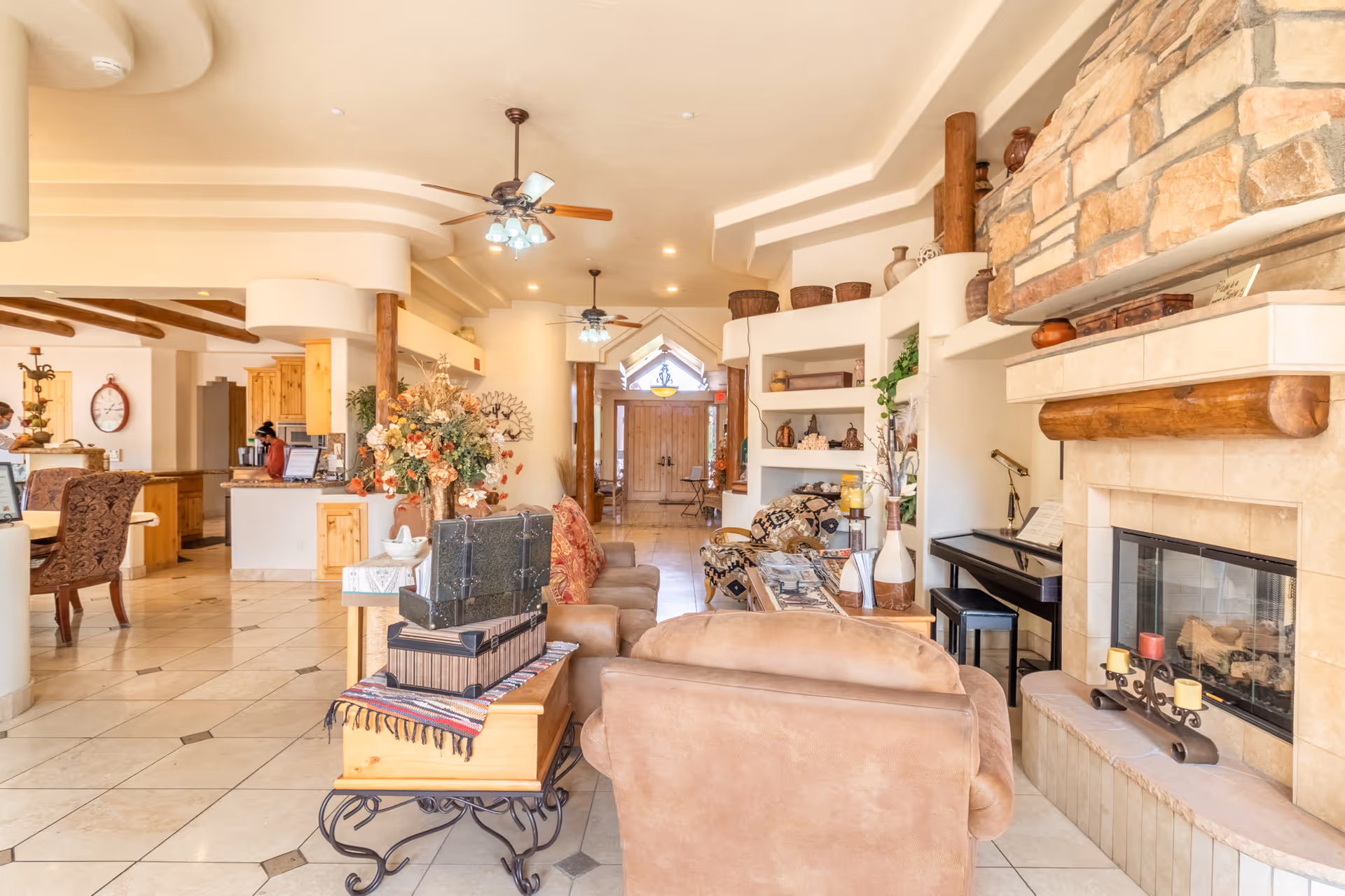 A spacious and warmly decorated living room area in Crismon Peaks Assisted Living featuring beige tiled floors, comfortable brown sofas, a stone fireplace with candles on the hearth, a piano, ceiling fans with lights, and decorative shelves with pottery and plants. The room opens into a dining area and kitchen with wooden cabinetry and a large clock on the wall.