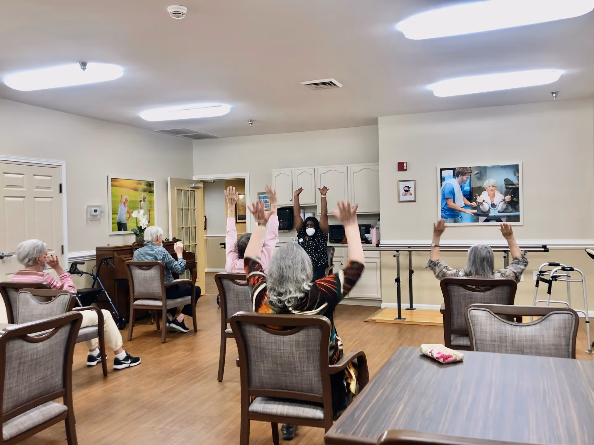 A group of elderly individuals seated in chairs in a well-lit room participating in a seated exercise session led by an instructor wearing a mask. The room has wooden flooring, light-colored walls, and framed pictures on the walls. Some participants have their arms raised during the exercise.