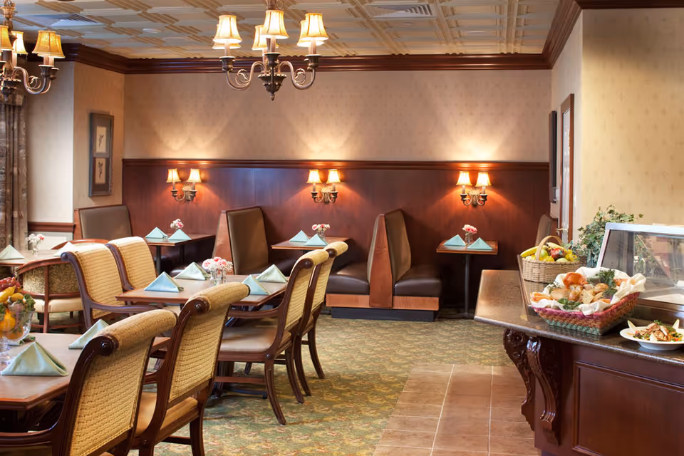 A cozy dining room in The Woodlands Retirement Community featuring wooden tables set with folded napkins and small flower arrangements. There are cushioned chairs and booth seating along the wall with warm wall sconces and chandeliers providing soft lighting. A buffet area with baskets of bread and fruit is visible on the right side.