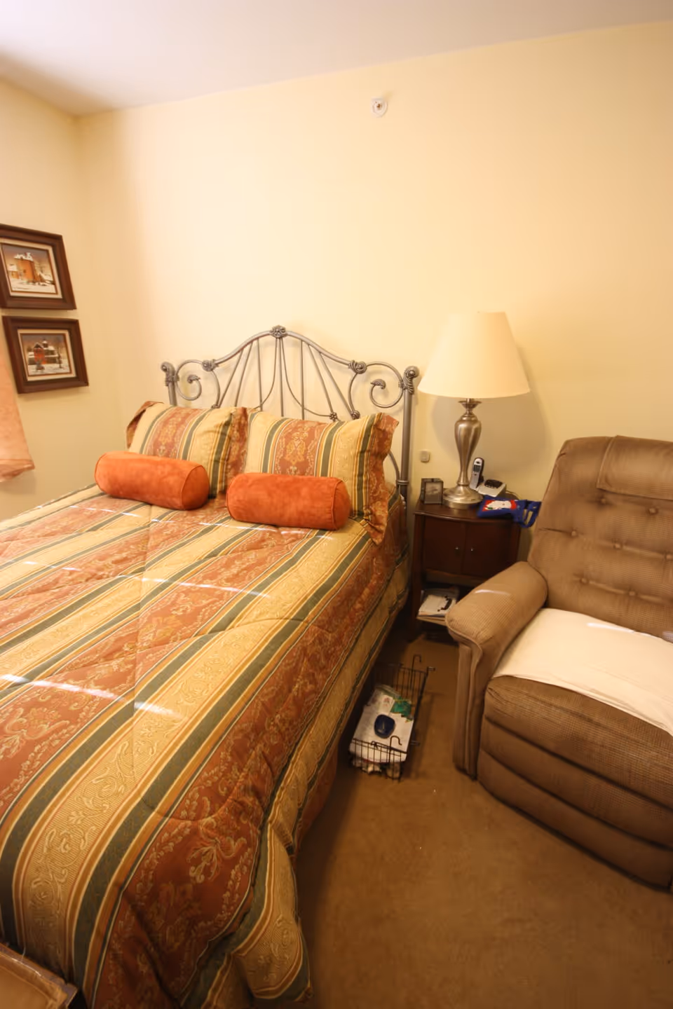 A cozy bedroom with a bed featuring a decorative metal headboard, striped and patterned bedding in shades of orange, green, and beige, and two cylindrical orange pillows. Next to the bed is a wooden nightstand with a silver lamp, a phone, and some small items. To the right of the nightstand is a brown upholstered recliner chair with a white cloth draped over the armrest. The walls are painted light yellow, and two framed pictures hang on the wall to the left.