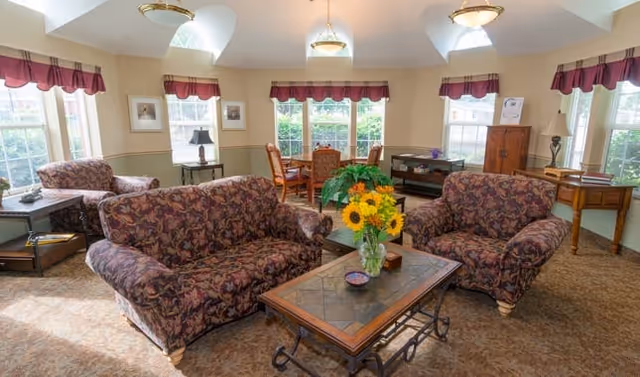 A bright and cozy living room area in a senior living facility with two patterned sofas facing a wooden coffee table adorned with a vase of sunflowers. The room has multiple windows with red valances, a round dining table with chairs in the background, and various wooden furniture pieces including side tables and a cabinet.