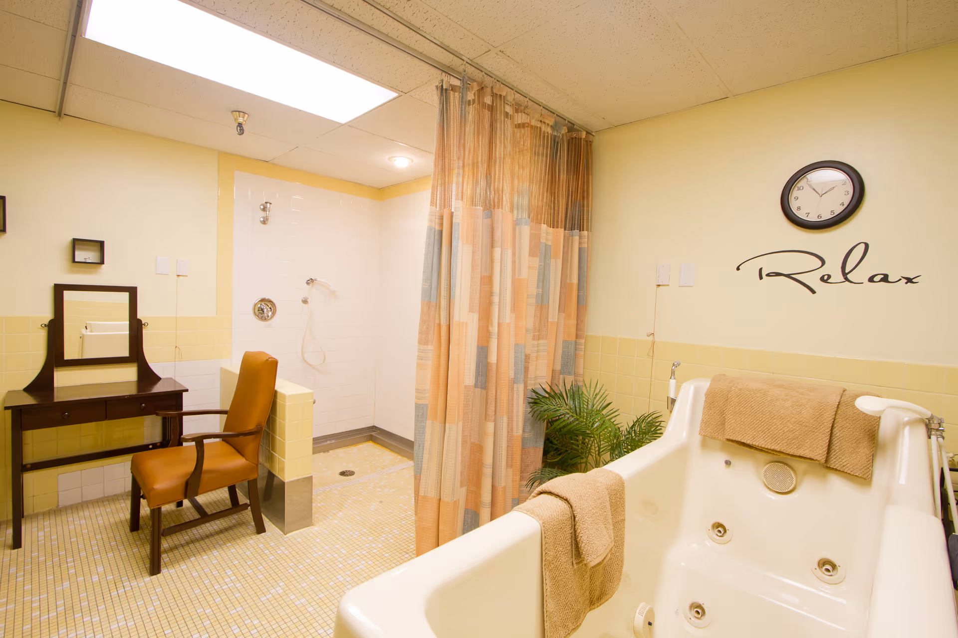 Bright bathing room with a white whirlpool tub, curtained walk-in shower, vanity and chair, and a 'Relax' wall clock.