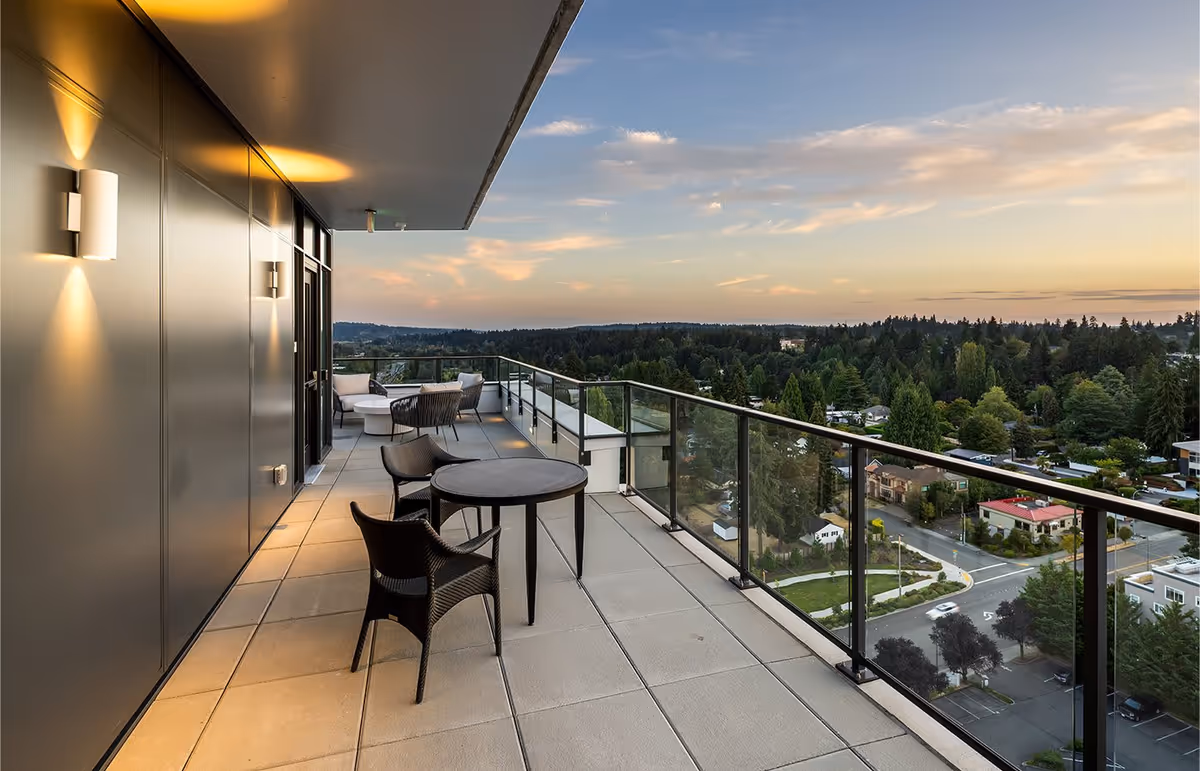 A spacious outdoor balcony with a glass railing overlooking a scenic view of trees, houses, and a road at sunset. The balcony has modern wall lights, a small round table with two chairs, and a seating area with cushioned chairs and a round coffee table.