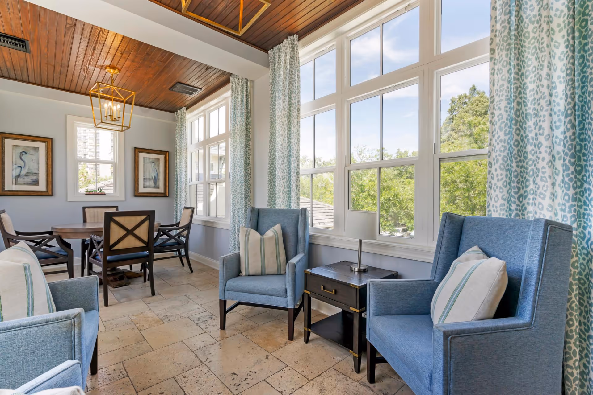 A bright and cozy sitting area with two blue armchairs with striped pillows, a small dark wooden side table with a lamp between them, and large windows with patterned curtains letting in natural light. In the background, there is a dining area with a round wooden table and four chairs, framed bird artwork on the walls, and a wooden ceiling with a modern gold chandelier.