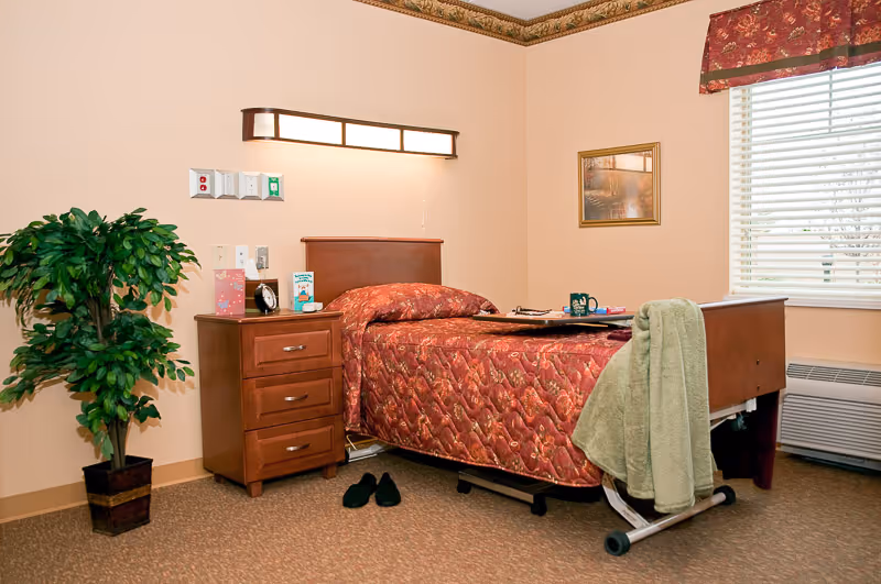 A single bed with a red patterned bedspread in a senior living facility room. Next to the bed is a wooden nightstand with drawers, a clock, and some books. A green towel is draped over the footboard of the bed. There is a potted plant on the floor to the left, a framed picture on the wall, and a window with blinds and a red valance on the right. The room has beige walls and carpeted floor.