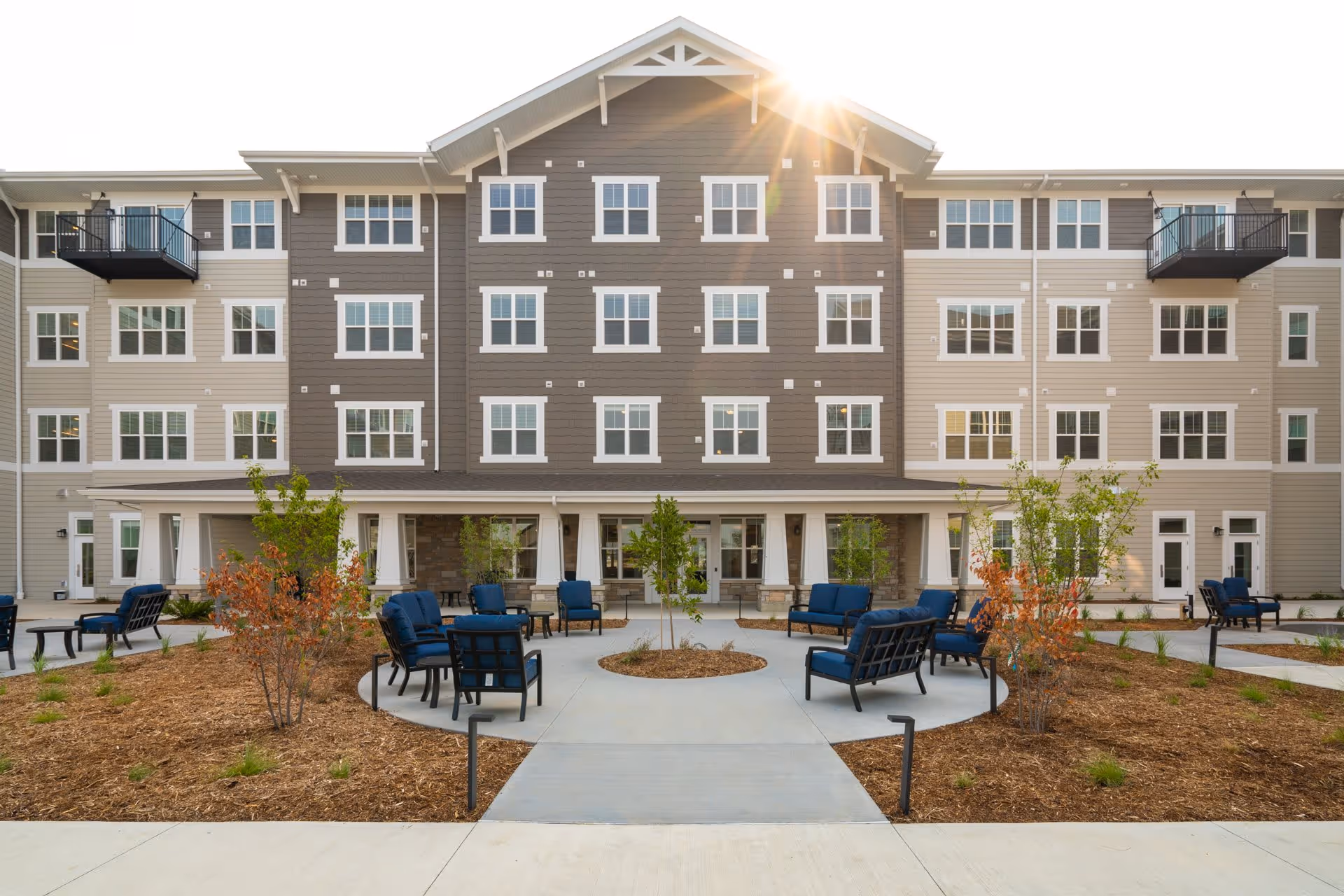 Exterior view of a multi-story senior living facility building with beige and gray siding, multiple windows, and small balconies. In front of the building is a landscaped outdoor seating area with blue cushioned chairs arranged around small tables and young trees planted in mulched beds. The sun is shining brightly above the building.