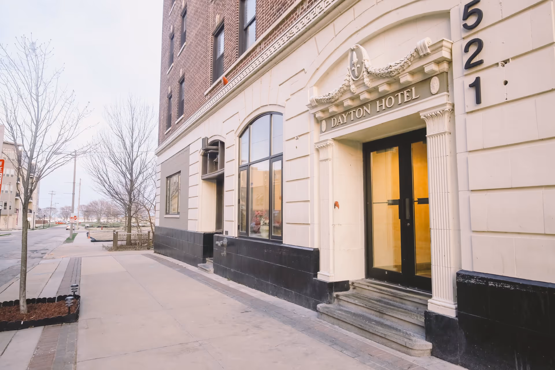 Exterior view of the Dayton Hotel building entrance with steps leading to glass double doors, beige and black facade, and the street sidewalk with leafless trees.