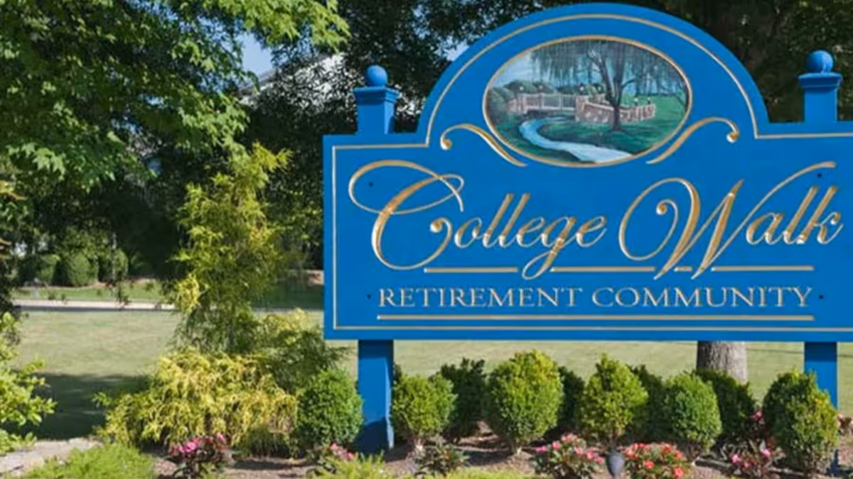 A blue sign with gold lettering that reads 'College Walk Retirement Community' surrounded by green bushes and trees in an outdoor setting.