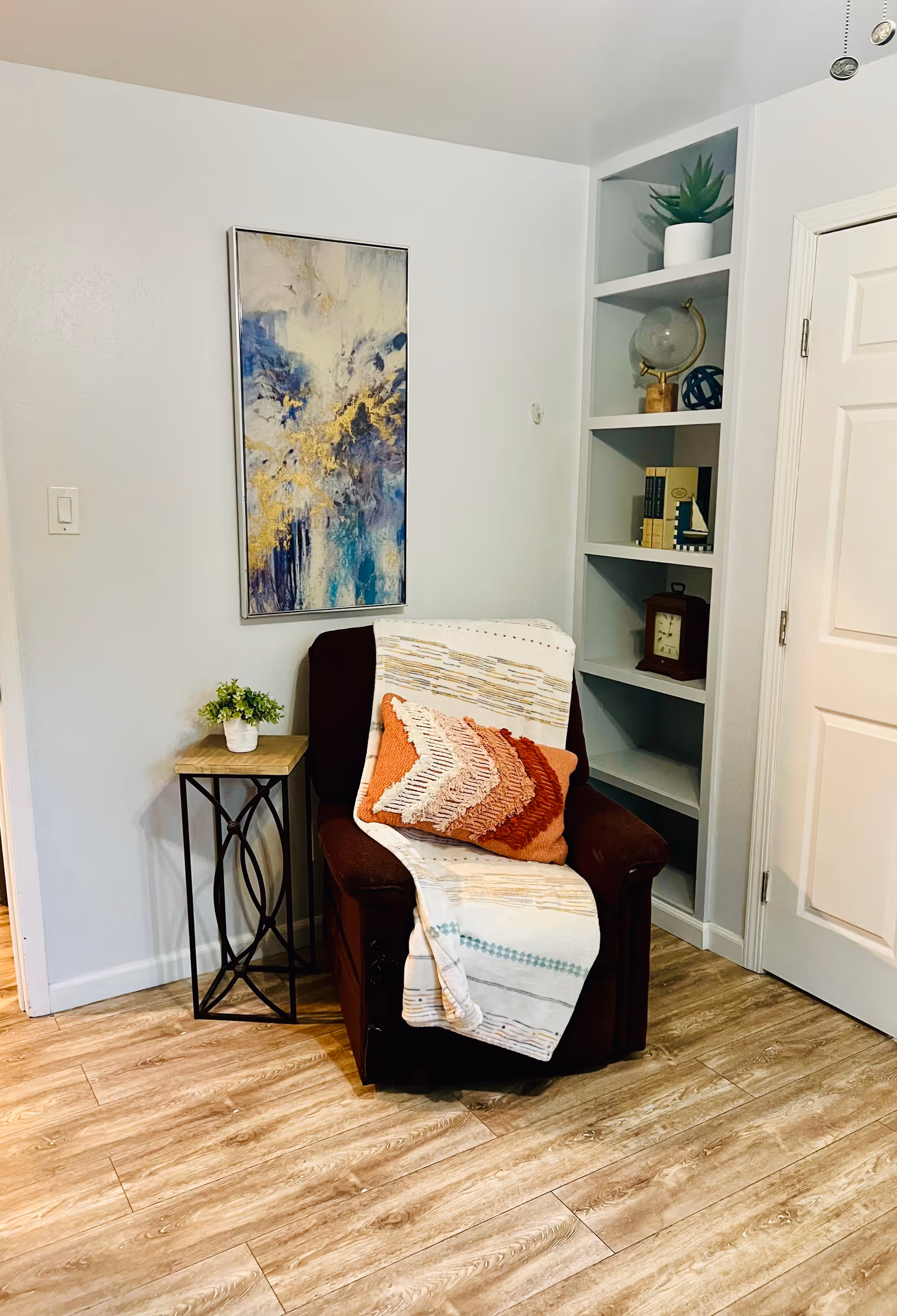 A cozy corner of a room featuring a dark brown armchair draped with a light-colored throw blanket and an orange textured pillow. Next to the chair is a small side table with a potted plant. On the wall above the chair hangs an abstract painting with blue, white, and gold tones. To the right, built-in shelves hold decorative items including a globe, books, and a clock. The floor is light wood, and a white door is visible on the right side.