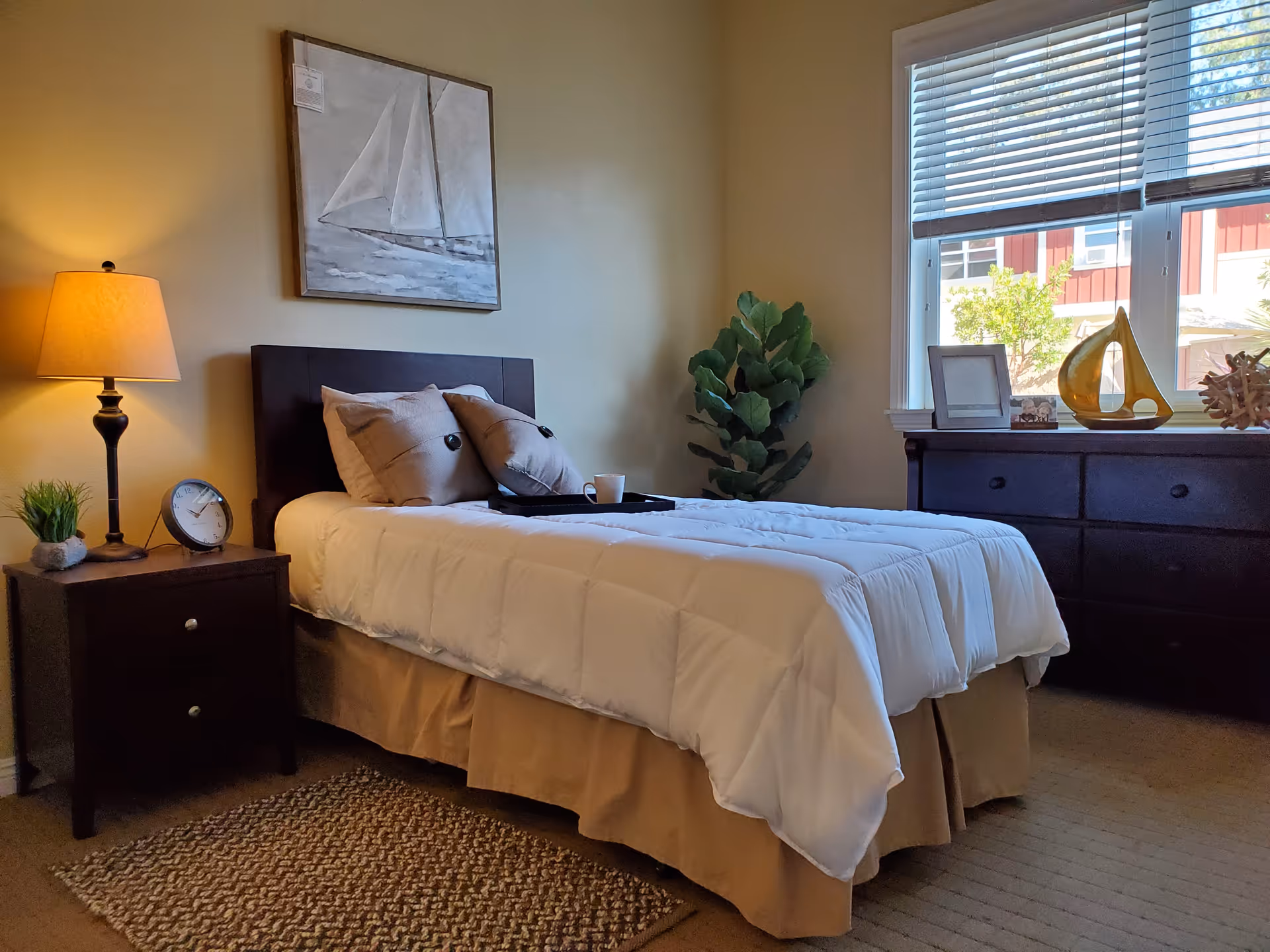 A neatly made single bed with white bedding and two beige pillows in a bedroom. Next to the bed is a dark wooden nightstand with a lamp, a small plant, and a clock. On the wall above the bed is a painting of a sailboat. A window with blinds lets in natural light, and below it is a dark wooden dresser with decorative items and a small plant beside it.