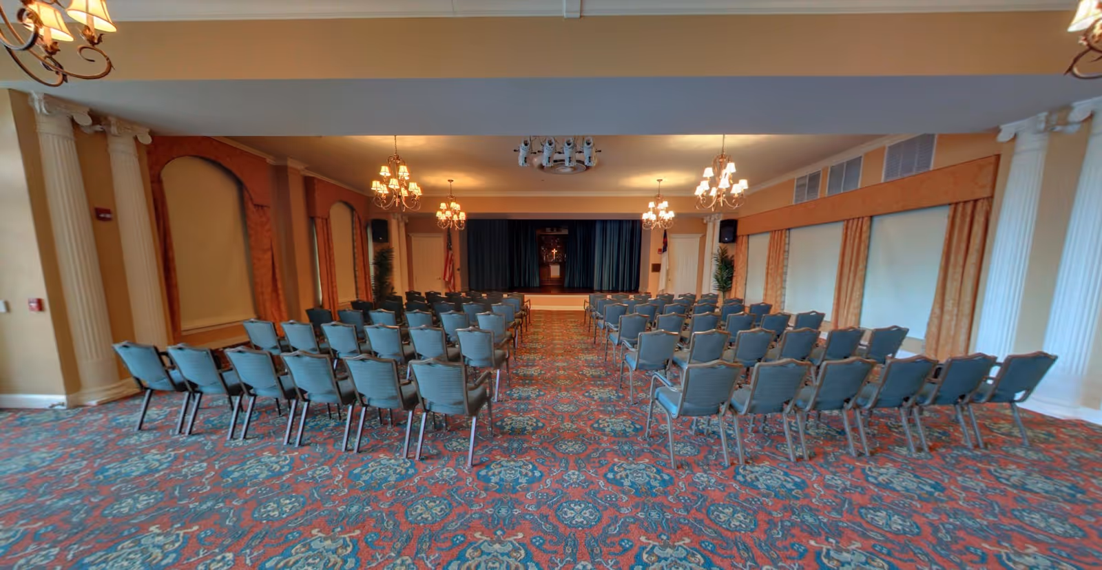 An empty event room with rows of chairs facing a small stage, chandeliers, ornate columns, and patterned carpet.