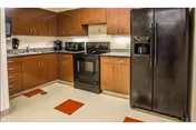 A kitchen area with wooden cabinets, a black refrigerator with a water dispenser, a black stove with an oven, a microwave, a coffee maker, and a toaster on the countertops. The floor has a light color with red square tiles interspersed.