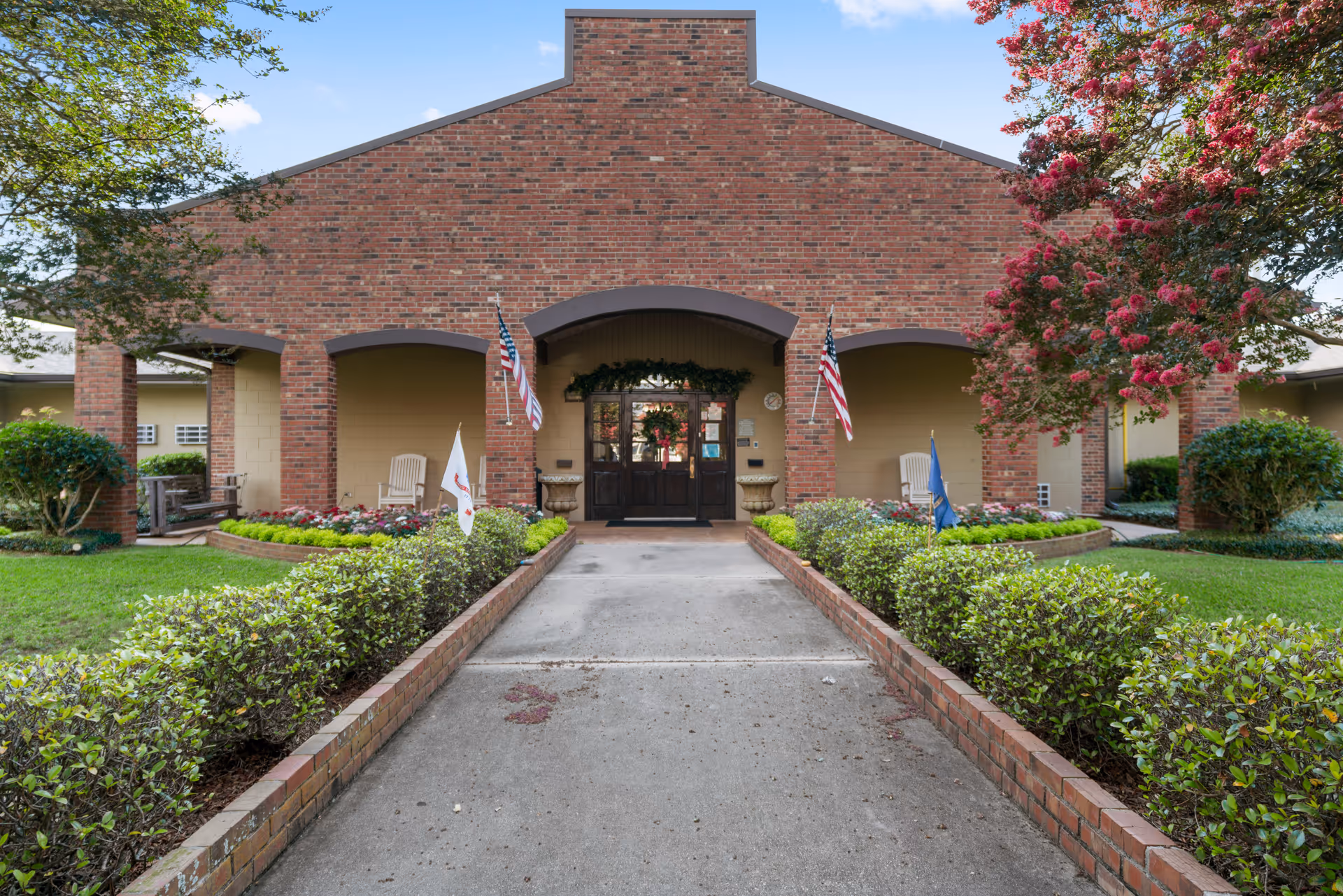Front entrance of a brick building with a concrete walkway bordered by low hedges leading to double wooden doors. The entrance is decorated with two American flags, flower beds, and greenery. There are chairs on the porch area under the arches, and trees with pink flowers flank the building.