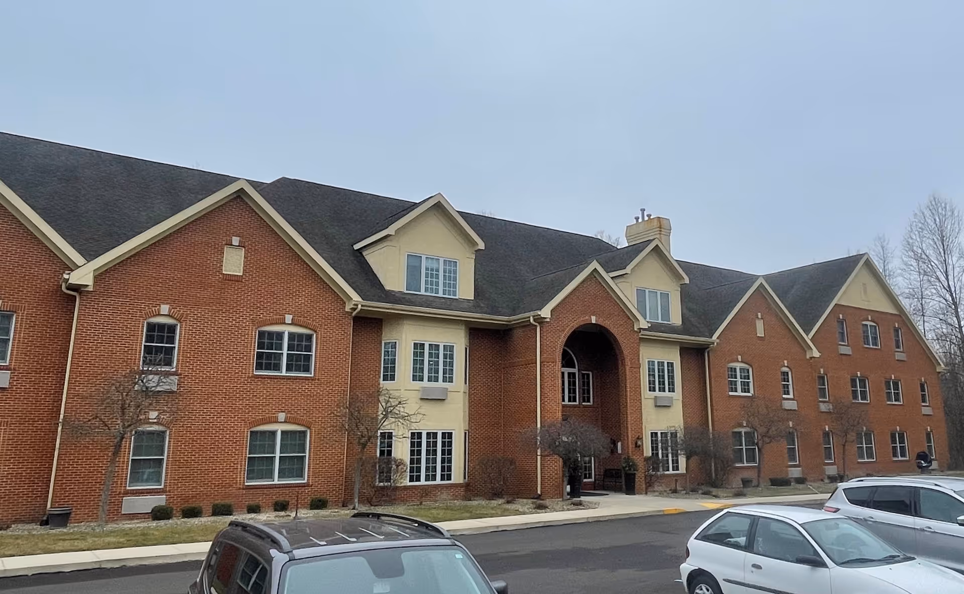 Exterior view of a two-story senior living facility building with red brick and beige accents, multiple windows, a large arched entrance, and several parked cars in front under a cloudy sky.