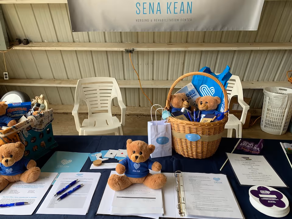 Table set up with informational materials, pens, and teddy bears wearing Sena Kean Nursing and Rehabilitation Center shirts. There is a wicker basket filled with more teddy bears and promotional items. Behind the table are white plastic chairs and a banner that reads 'Sena Kean Nursing & Rehabilitation Center'.