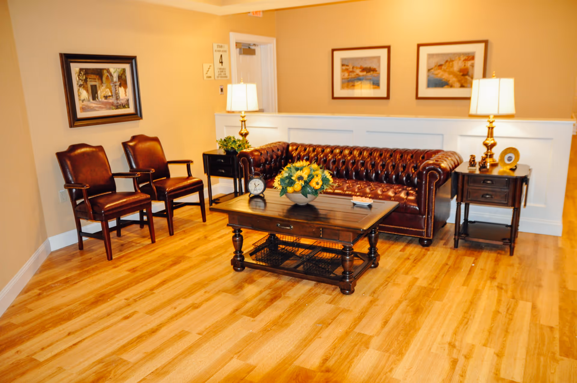 A cozy living room area with a dark brown leather tufted sofa, two matching leather chairs, a wooden coffee table with a flower arrangement and a clock on it, two side tables with lamps, and framed artwork on the walls. The floor is light wood, and the walls are painted beige.