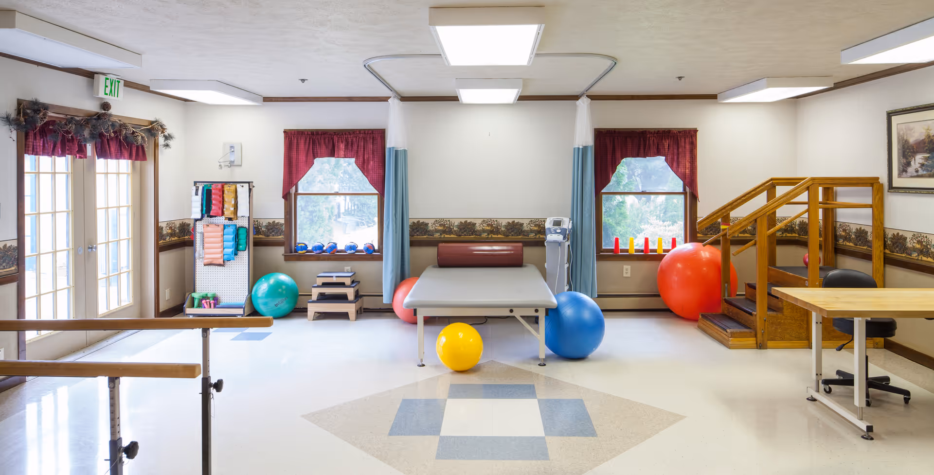 Bright interior therapy room with a treatment table, exercise balls, parallel bars and various rehab equipment.