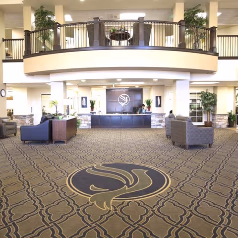 Spacious and well-lit lobby area of a senior living facility with patterned carpet featuring a large decorative emblem in the center. The reception desk is straight ahead with two staff members behind it, flanked by plants and wall art. Comfortable seating areas with sofas and chairs are arranged on either side. An upper balcony with railing overlooks the lobby, decorated with plants and a table with chairs.