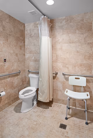 Accessible bathroom with beige tiled walls and floor, featuring a toilet with grab bars on both sides, a shower area with a white curtain, and a white shower chair with metal legs.
