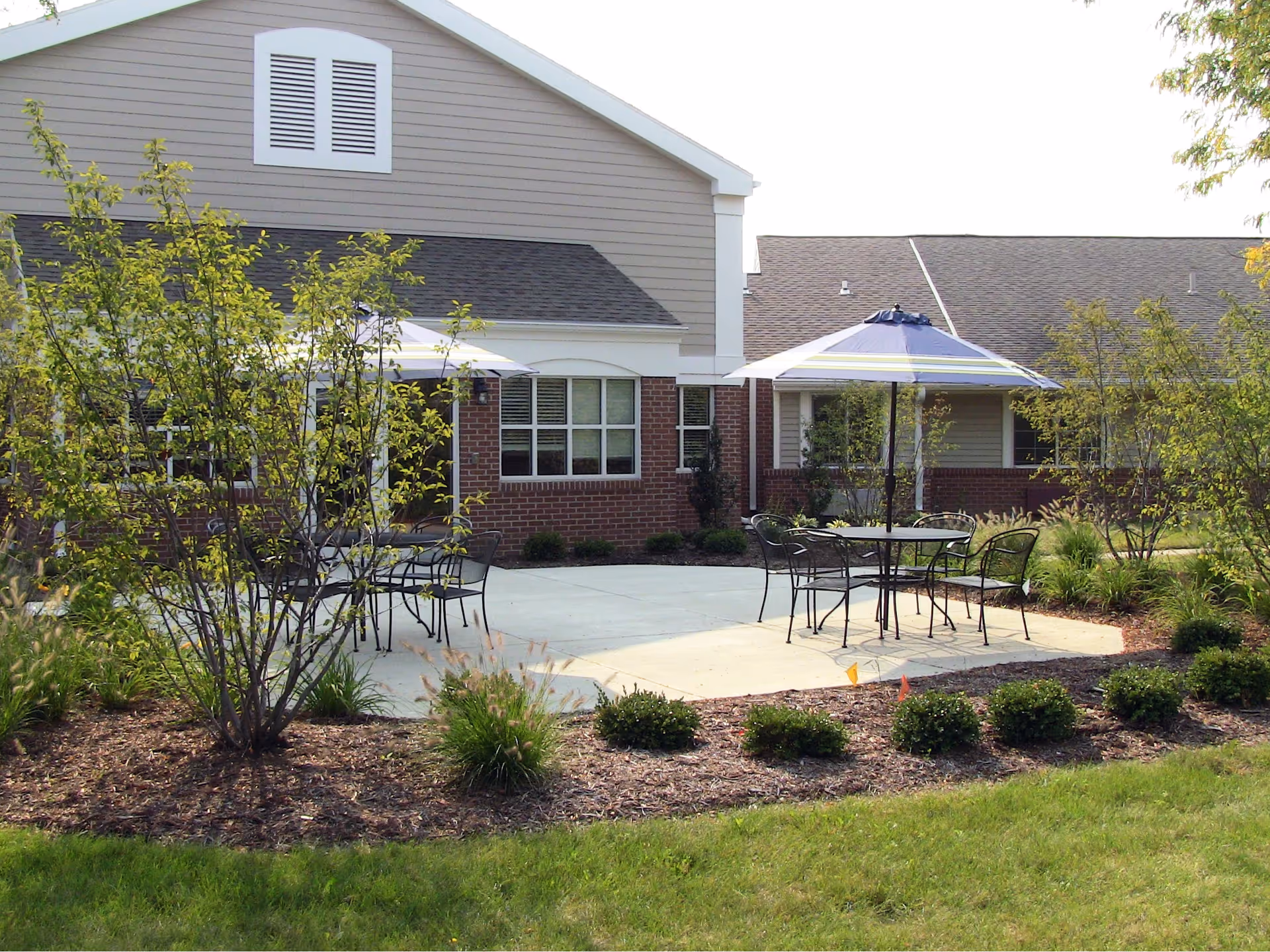Outdoor patio area with metal tables and chairs under umbrellas, surrounded by landscaped bushes and trees, adjacent to a building with brick and siding exterior.