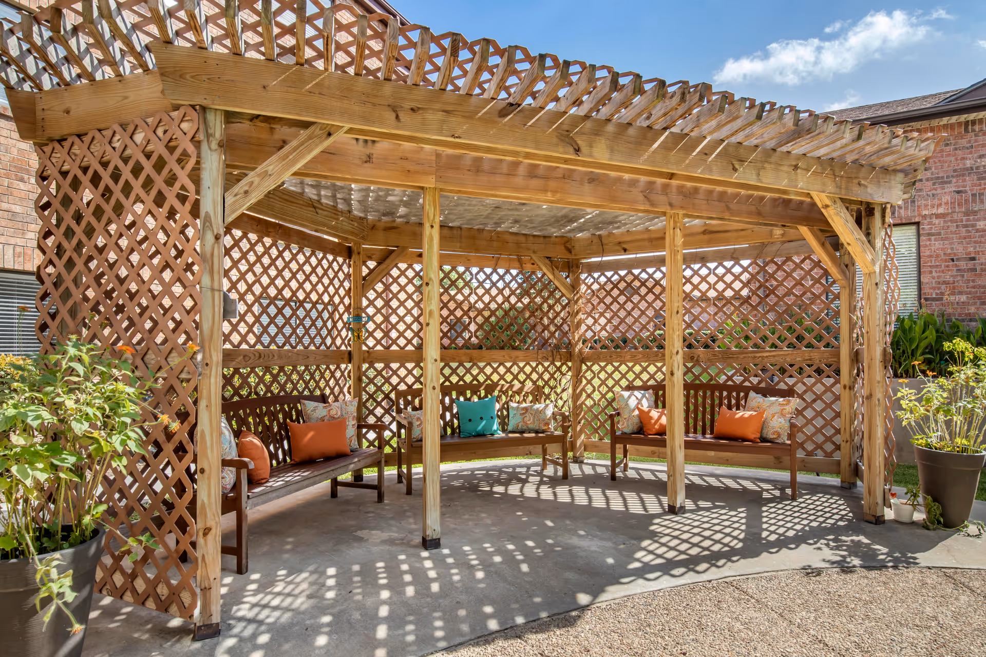 Wooden pergola with lattice walls and curved benches topped with colorful pillows in an outdoor courtyard.