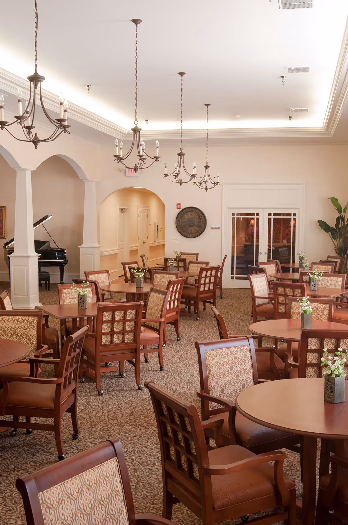 Spacious dining room with round wooden tables, upholstered chairs, chandeliers, and a piano in the background.