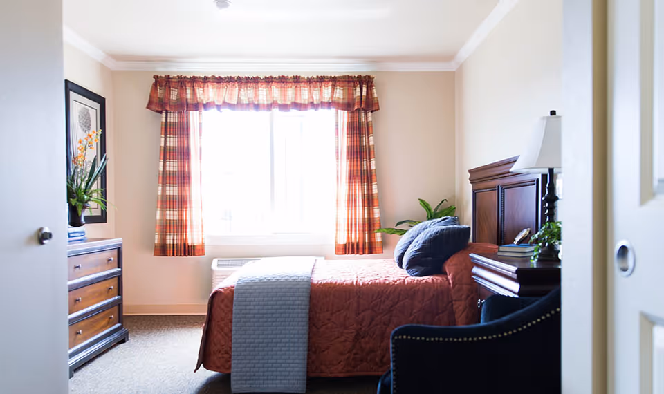 A cozy bedroom with a bed covered in a rust-colored quilt and a gray throw blanket. The bed has two dark blue pillows and a wooden headboard. Next to the bed is a wooden nightstand with a white lamp, books, and a small plant. Across from the bed is a wooden dresser with a framed picture and a vase with flowers on top. The room has a window with red and beige plaid curtains letting in natural light. A dark blue upholstered chair is partially visible in the foreground.