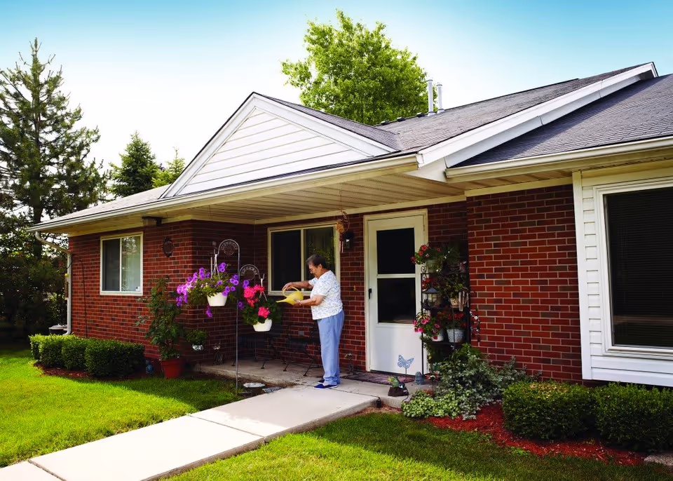 An elderly woman watering hanging flower pots on the porch of a single-story brick house with a white door and windows, surrounded by green grass and shrubs under a clear sky.