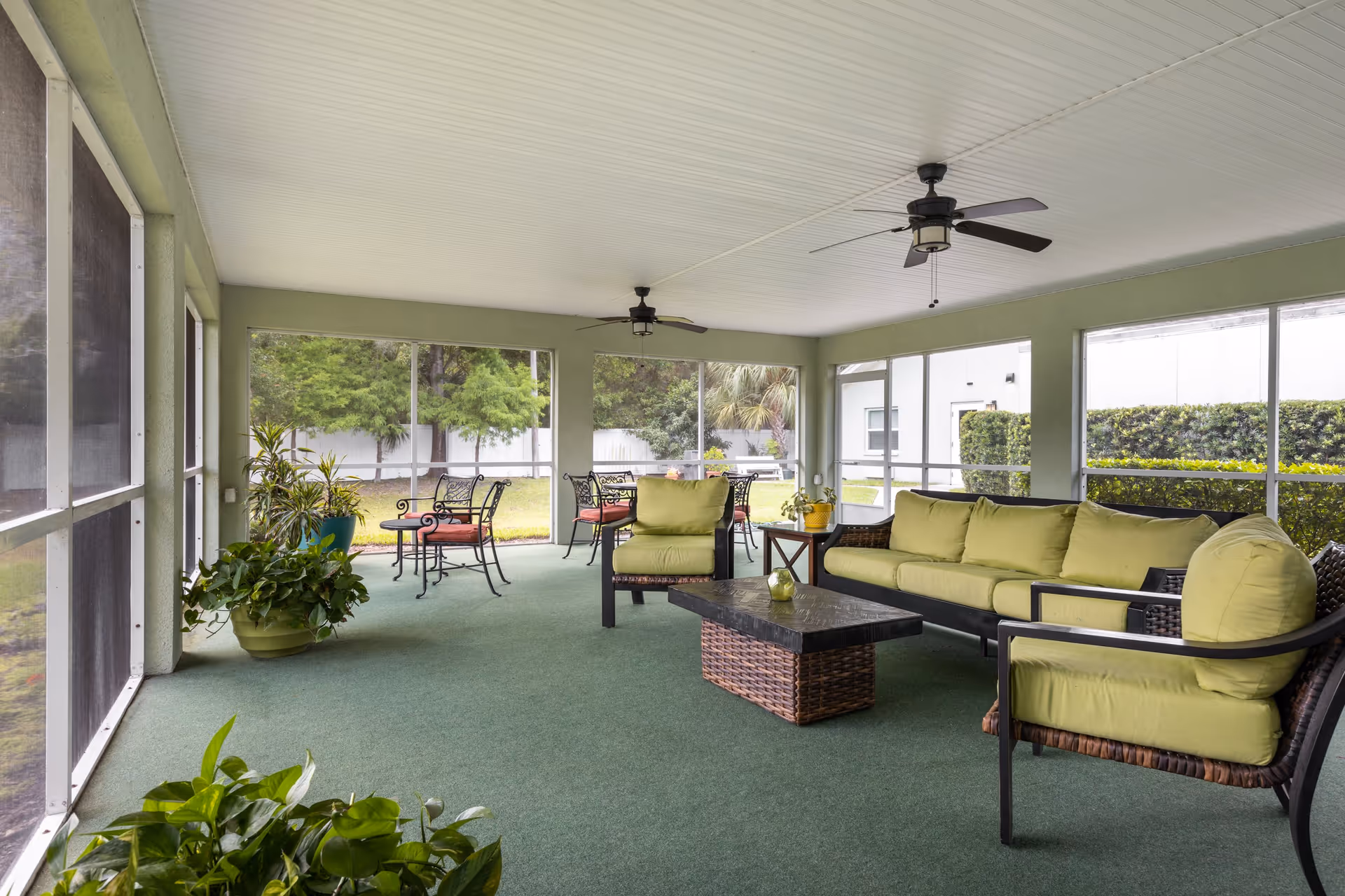 Screened-in patio with green-cushioned wicker seating, a coffee table, dining set, potted plants, and ceiling fans overlooking a lawn.