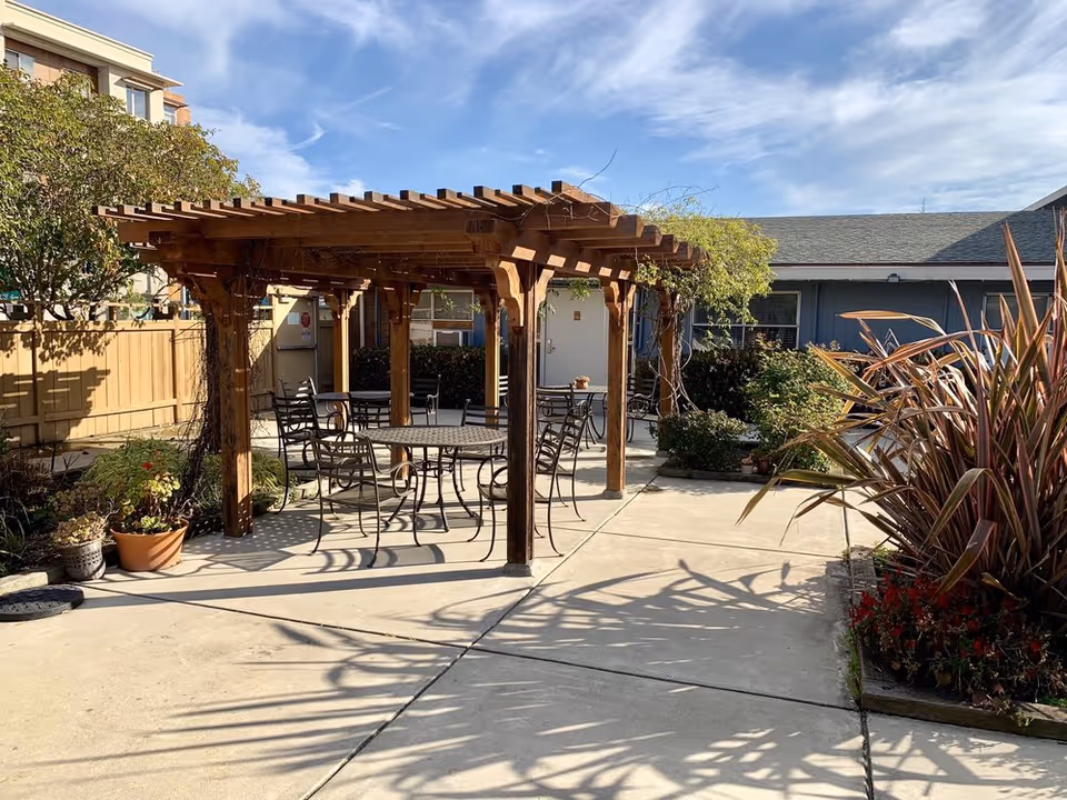 Outdoor patio area with a wooden pergola providing partial shade over several metal tables and chairs. Surrounding the patio are various plants and shrubs, with a wooden fence and building visible in the background under a partly cloudy sky.