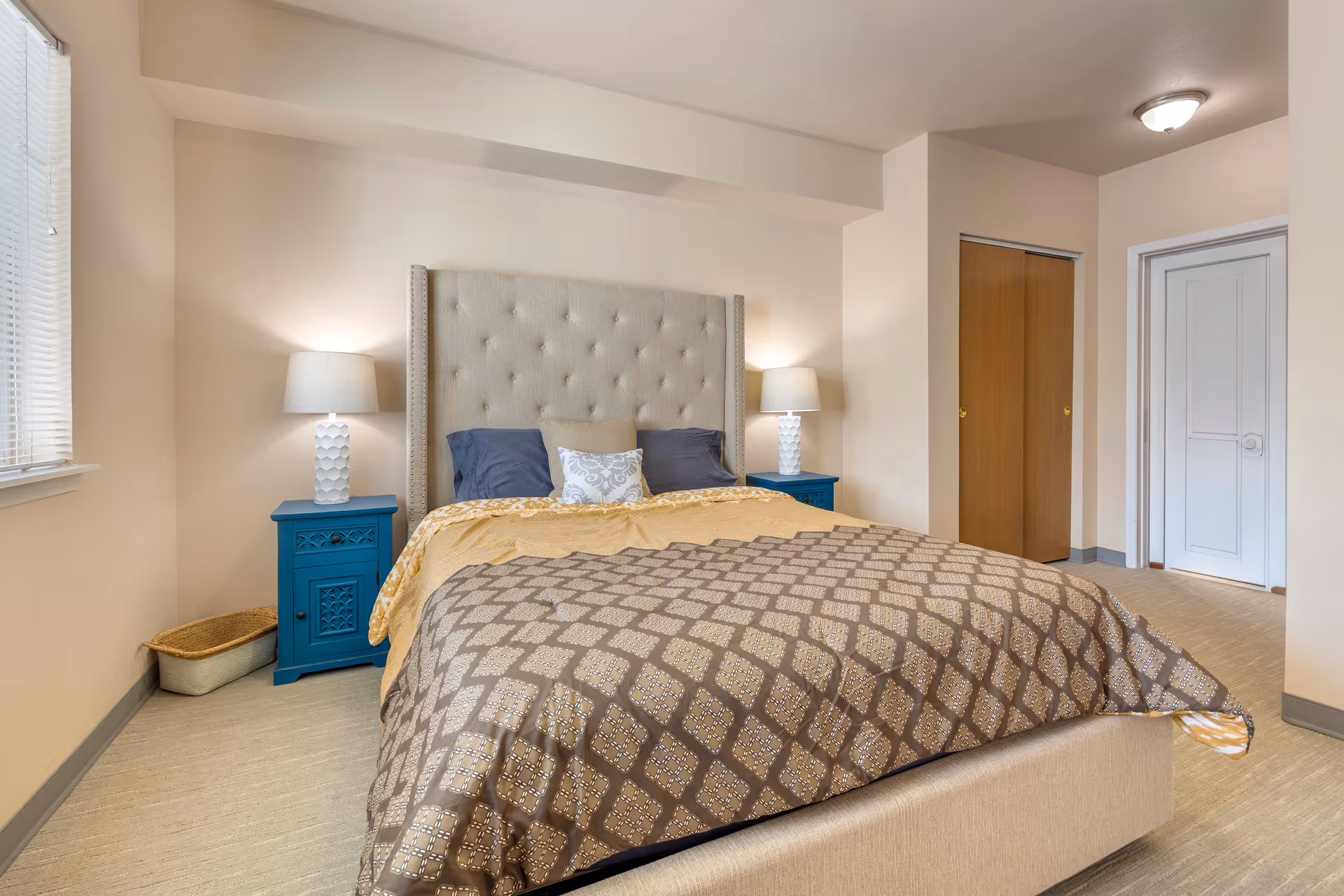 A bedroom with a large beige upholstered bed featuring a tufted headboard. The bed is made with a patterned brown and beige comforter, yellow blanket, and several pillows. On each side of the bed are blue nightstands with white lamps. There is a window with blinds on the left side, a wicker basket on the floor, and a closet with wooden sliding doors on the right side. The walls are painted light beige and the floor is carpeted.