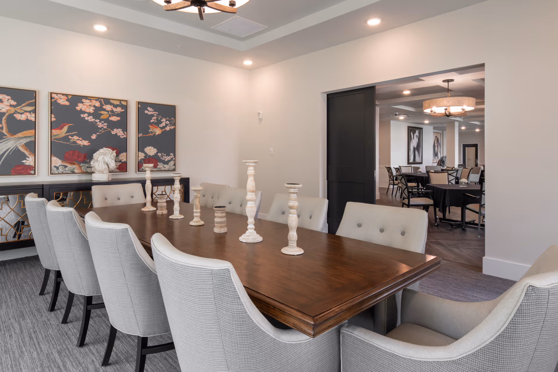 A modern dining room with a long wooden table surrounded by cushioned chairs. The table is decorated with various white candle holders. On the wall behind the table, there are three framed artworks featuring birds and floral designs. In the background, another dining area with tables and chairs is visible through a wide doorway.