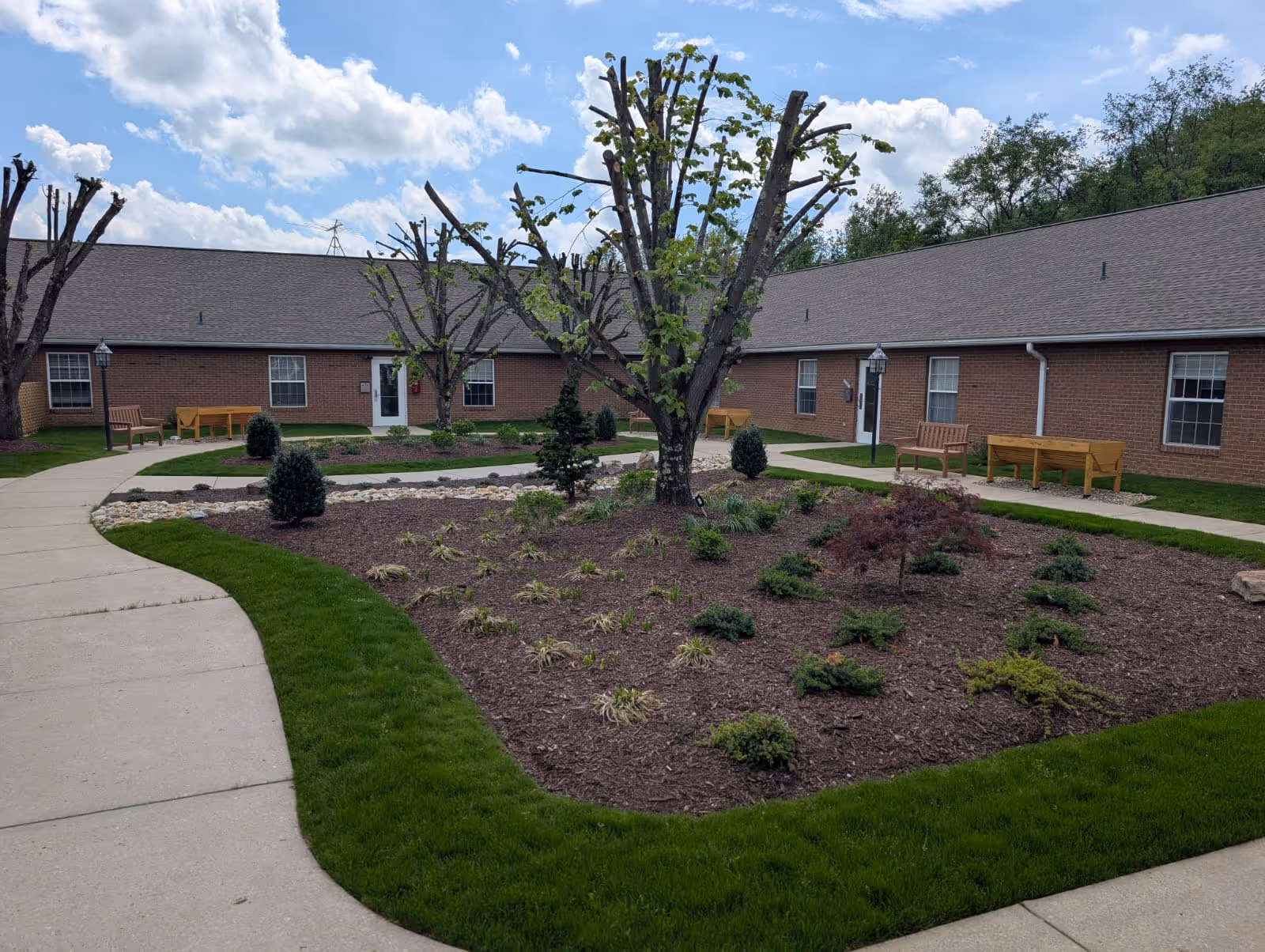 Brick single-story buildings surrounding a landscaped courtyard with trimmed trees, benches and winding concrete walkways under a partly cloudy sky.
