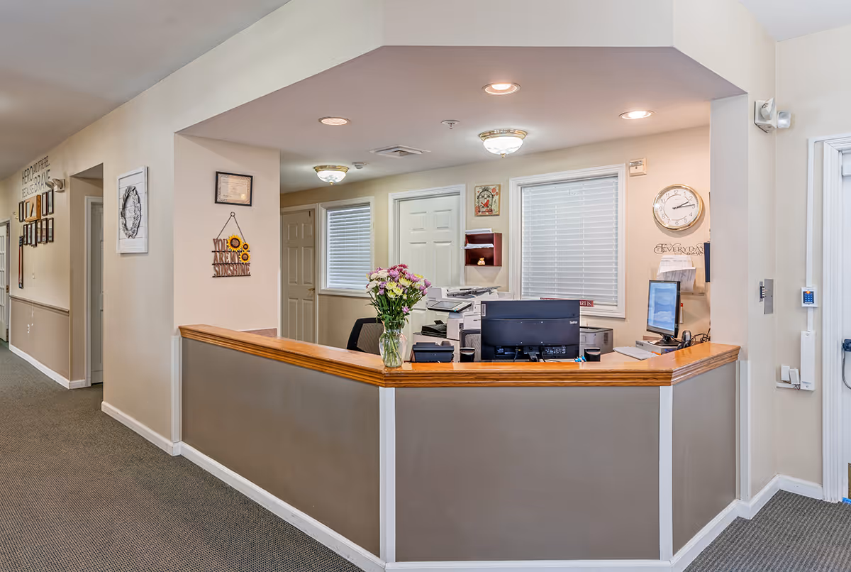 Reception desk area inside Floyd Senior Living facility with a wooden countertop, computer monitors, a printer, and a vase of flowers. The walls are light-colored with framed decorations and a clock. There is a hallway to the left with additional wall decorations and doors behind the desk.