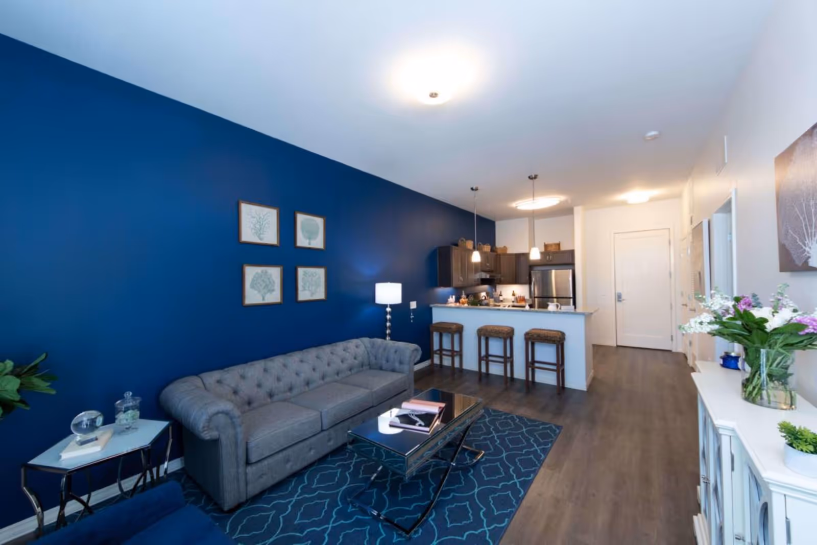 A modern senior living apartment interior featuring a gray tufted sofa against a blue accent wall with four framed botanical prints. A glass coffee table sits on a patterned blue rug in front of the sofa. To the right is a kitchen area with a breakfast bar, three bar stools, dark cabinets, and stainless steel appliances. The floor is a dark wood laminate, and there are decorative plants and flowers on a white cabinet along the right wall.