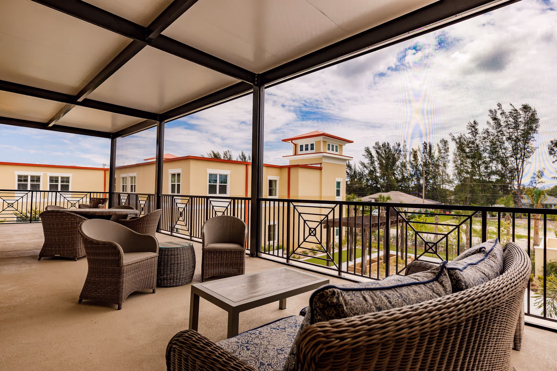 Covered outdoor patio area with wicker chairs, a wicker sofa with patterned cushions, a small coffee table, and a round wicker side table. The patio overlooks a landscaped garden area with trees and a beige building with red trim in the background under a partly cloudy sky.