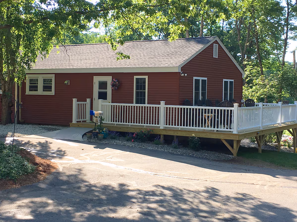 A single-story red building with white trim and a gray shingled roof surrounded by trees. The building has a white railing deck with several black chairs and a small table. There is a paved driveway in front and some potted plants near the entrance.