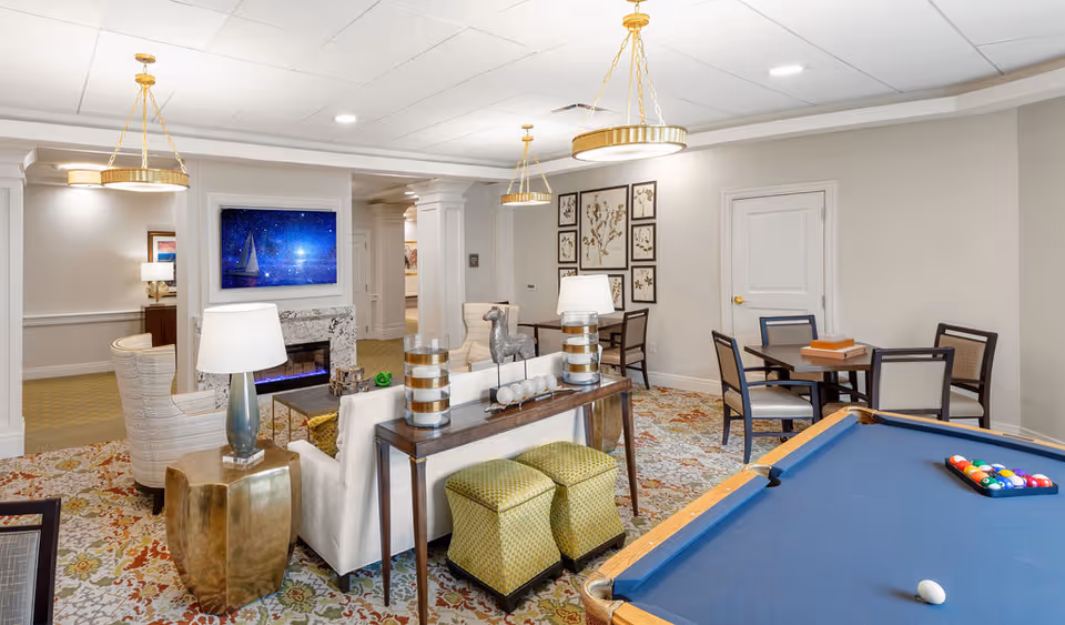 A well-lit common area in a senior living facility featuring a pool table with balls set up, a seating area with a sofa, armchairs, side tables with lamps, a fireplace with a framed picture above it, and a dining table with chairs. The room has patterned carpet, neutral-colored walls, and decorative wall art.