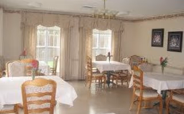 A dining room with several wooden tables and chairs. Each table is covered with a white tablecloth and has a small vase with flowers. There are large windows with beige curtains allowing natural light to enter the room. Two framed pictures hang on the wall.