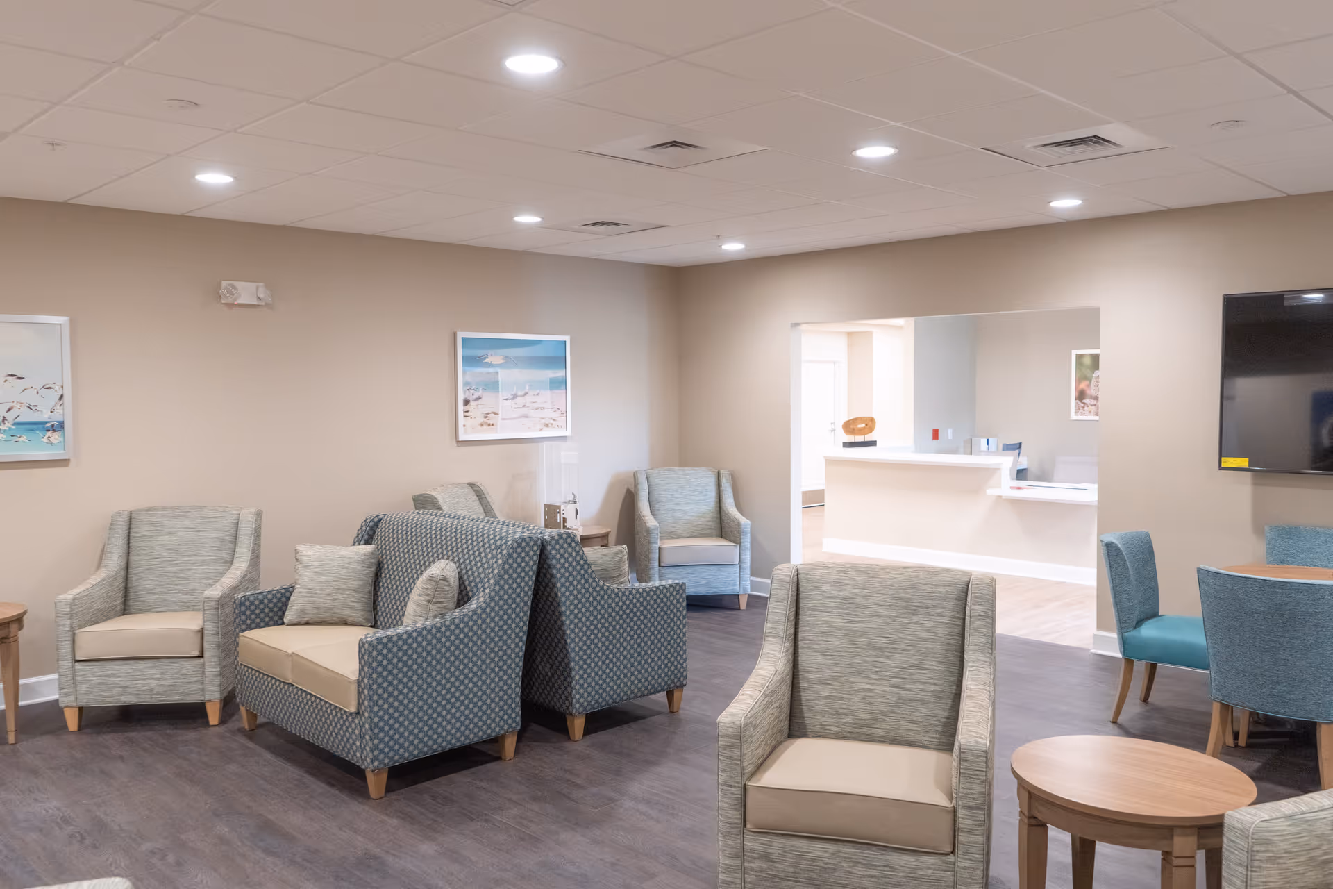 A well-lit senior living facility common area with several upholstered armchairs and loveseats arranged around small wooden tables. The walls are beige with framed beach-themed artwork. In the background, there is a reception desk area and a mounted flat-screen TV on the right wall.