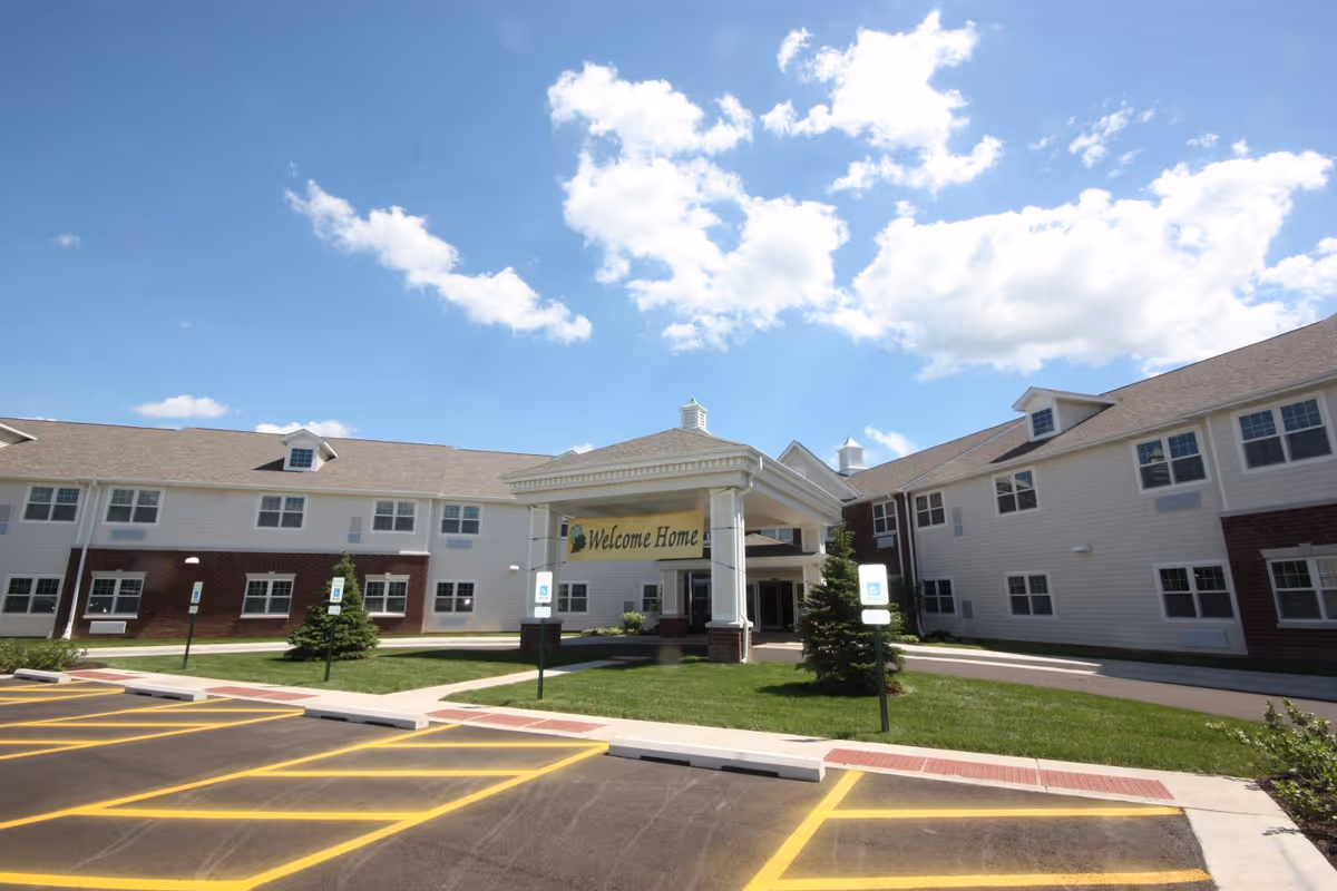 Exterior view of a senior living facility building with a covered entrance displaying a 'Welcome Home' sign. The building has two stories with white siding and brick accents, surrounded by a parking lot with marked spaces and a clear blue sky with scattered clouds above.