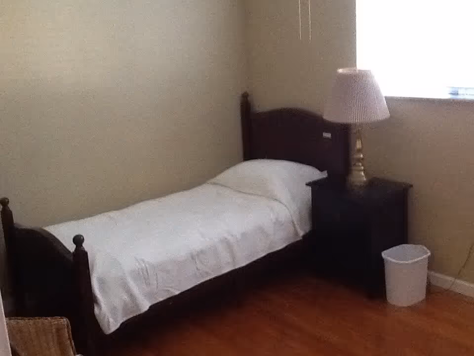 A simple bedroom with a single wooden bed covered with a white sheet and pillow. Next to the bed is a dark wooden nightstand with a brass lamp and a white lampshade. A small white trash bin is on the floor beside the nightstand. The room has light-colored walls and a window letting in natural light.