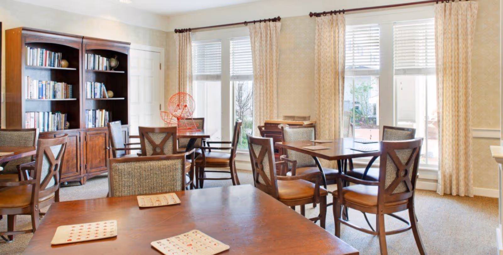 A bright room with multiple wooden tables and chairs arranged for group activities. There is a large wooden bookshelf filled with books and decorative globes against the wall. The room has large windows with white blinds and patterned curtains, letting in natural light. On one table, there is a red bingo cage, and bingo cards are placed on the tables.
