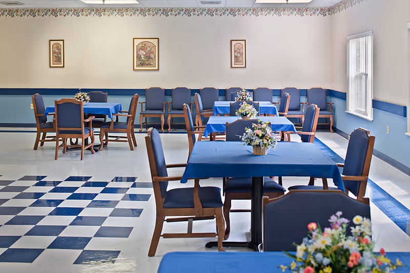 A dining room with several tables covered with blue tablecloths, each adorned with a floral centerpiece. Wooden chairs with blue cushions surround the tables. The room has a checkered blue and white floor pattern in the foreground, light-colored walls with floral wallpaper border near the ceiling, and framed floral artwork on the walls. There are windows on the right side letting in natural light.